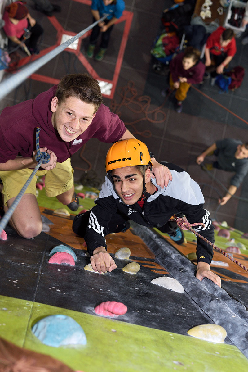 A climber ascending an indoor climbing wall assisted by one of our volunteers.