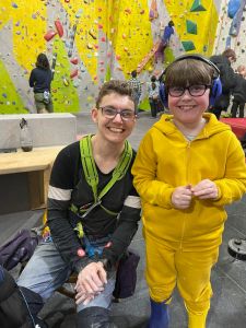 A person in climbing harness is kneeling on the floor next to a girl with headphones, both of them with big smiles