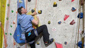 Smiling acceptingly, a confident climber sidepulls on a yellow volume, looking upwards for the next move