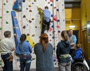 Cheerfully watched by 6 people standing or sat in wheelchair, a confident climber executes a move on climbing wall