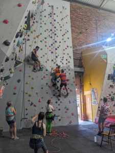 A long shot of 7 people, including climber and her side-climber plus a climber with camera on the climbing wall, and 4 people below operating ropes