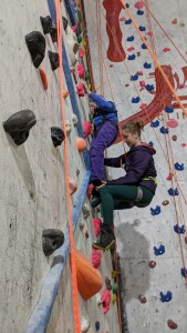 Side-on view of two women on climbing wall shows one climber focused intently on holdling on while climber behind guides her feet into position