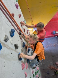 Looking down on two climbers, the woman doubly-roped and focusing on finding holds, the man side-climbing to assist