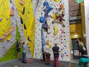 Two women are ascending a climbing wall, one with a side-climber giving assistance, with 5 people on the ground belaying or assisting with pulley ropes
