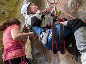 A smiling climber in parapente harness looks up at climbing wall as she ascends, with another woman side-climbing behind
