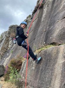 A climber in harness and helment is smiling as she abseils down a gritstone slab in Peak District