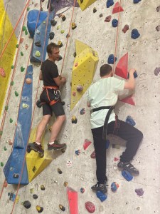 A man focuses on hand- and foot-holds ascending a climbing wall with a nearby climber, also roped, offering advice