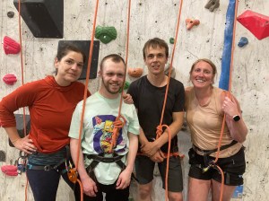 4 people are lined up at the foot of climbing wall, still attached to climbing ropes, smiling after a successful climb