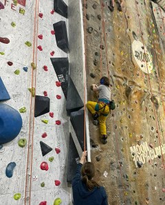 A girl pulls on a climbing hold with both hands as she steps up to next foothold, while a woman below holds the belay rope