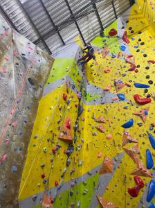 A lone climber is seen at the top of a high overhanging climbing wall, looking down
