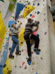 Roped climbers ascend a climbing wall, with smiling woman suggesting next move to man