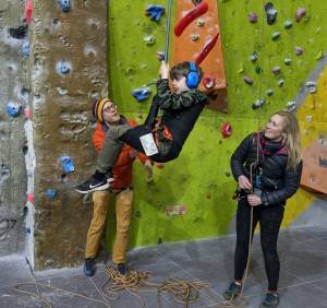 A boy in headphones sits in a parapente harness a metre above the ground, swinging gently as he is pushed by 2 volunteers, one of them holding safety rope