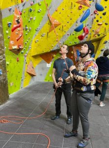 A young man wearing gaming-themed hoody looks up at climbing wall and lets rope through a belay device, while another man also looks up and holds end of rope