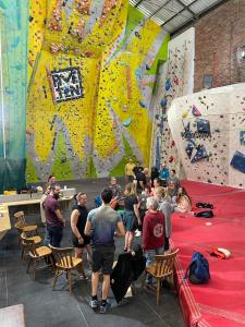 An informal group of around 18 people are stood or sitting on chairs or matting in a climbing hall