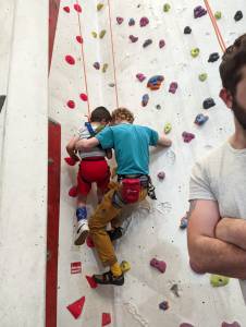 A young boy in a full body harness is ascending a corner route, supported by a man, also roped, with another volunteer half-seen in foreground