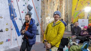 2 men in duvet jackets cheerfully operate belay ropes while a girl and 2 adults are sat on wooden lockers behind and spectating