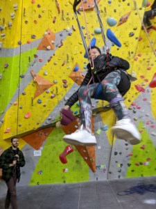 Bobble hat in her hand and sneakers kicking towards the camera, a girl sat in a parapente harness grins as she swings high in the air, while a smiling man operates safety rope in background