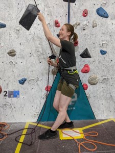 A smiling woman leans back and pulls on belay rope, climbing wall behind