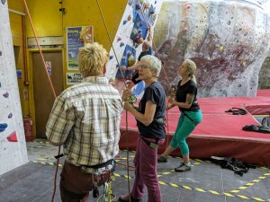 3 people stood in front of climbing wall are operating ropes via belay devices, and exchanging comments or looking up at wall