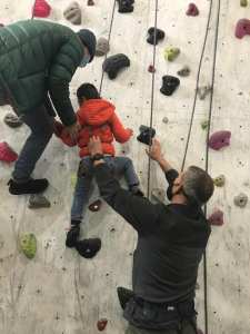 A boy reaches for climbing holds, helped by 2 people in covid-era masks, also roped and one beside the boy on the wall