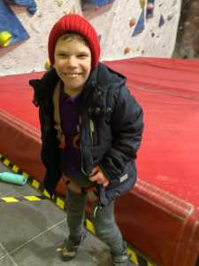 A boy in anorak and wooly hat, still wearing climbing harness, is standing and grinning broadly