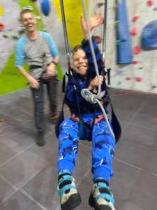 A smiling boy in parapente harness swings towards the camera, after the roped man stood behind has given him a push