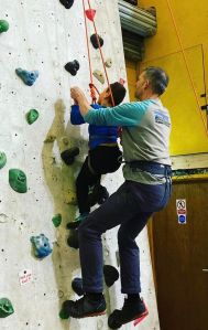 Both roped and part-way up climbing wall, a man helps a boy reach for climbing holds
