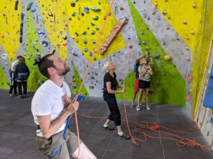 4 people look up at an unseen climber , a woman clasping hands in delight, a man aiming phone for photo, and 2 others operating ropes