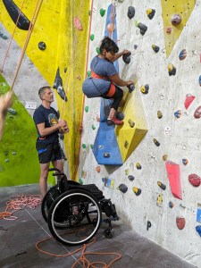 A climber with limb difference works her way up climbing wall, leaving wheelchair below on ground, while 2 people (one out of shot) operate ropes