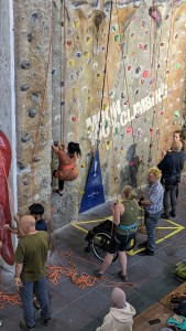 High-level long shot shows person working her way up wall, with wheelchair on ground below, and people holding belay rope and pulley rope