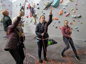 Team of three belayers operate ropes for unseen climbers above on the wall. Other people are climbing the wall behind.