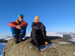 Under clear blue skies, two people in warm and rugged hiking clothes perch on rocky mountain top, with extensive snowy moorland seen below