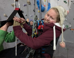 A climber in a paragliding harness smiles at the camera, with a sling attached by a carabiner and belaying line. Her gloved hands are supported on a bar by an unseen helper, and a belayer is operating the rope behind