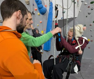 A climber suspended in a paragliding harness grasps a bar attached to pulley system, while Gordon belays, David operates the pulley, and Nick helps Clare’s hands to grip the bar