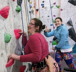 A smiling climber grips handholds while her belayer standing beside gives a thumbs up, and an unseen side supporter gives her harness a steadying hand