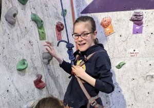 Alice is stood by the wall, tied in and ready to climb, with her hand on a hold as she looks round to smile at the camera.