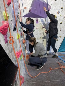 Alice reaches for hand holds, with volunteer Noni ready to guide her feet to holds, and Csilla belaying her.