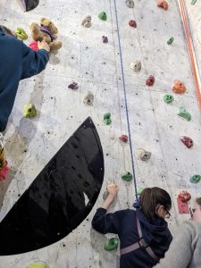 Caramel The Bear is perched on a climbing hold, observing Alice's climb.