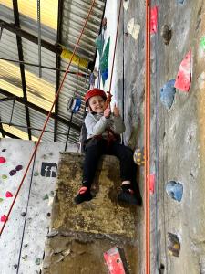 A double thumbs up from a young climber, who is sitting on the pillar part-way up the wall and smiling down at the camera.