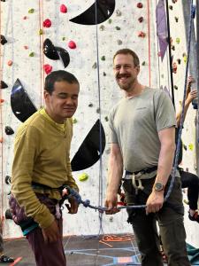 Ryan is roped up and ready to climb, stood near the climbing wall with volunteer Andrew B, who is smiling and ready to belay. 