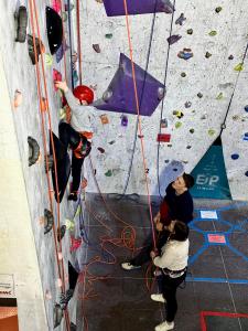A boy in a red helmet pulls himself up the climbing wall, with Robin backing up a family member who is learning how to belay.